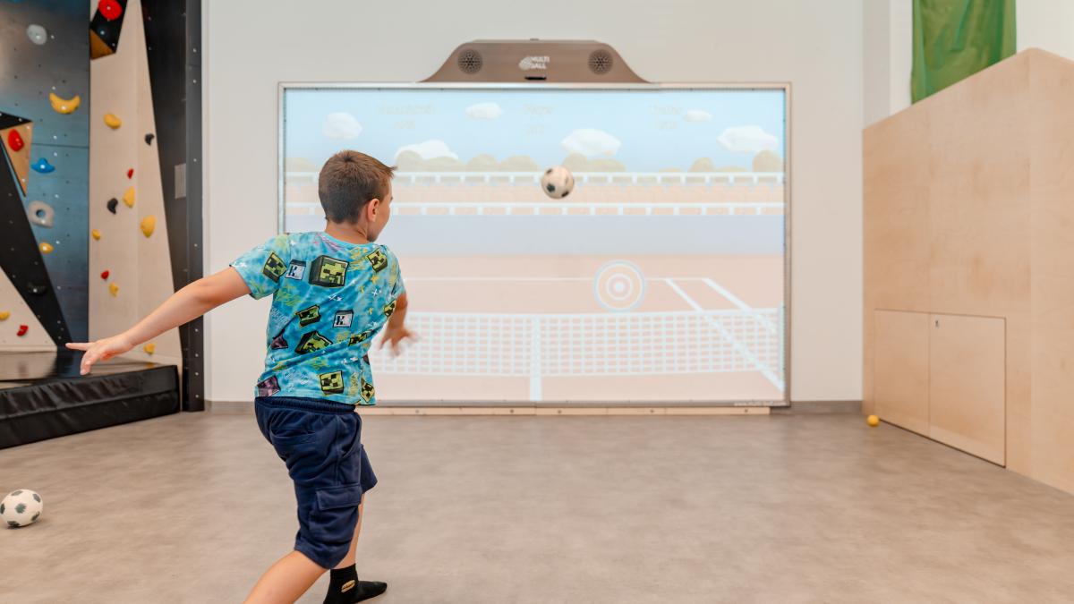 Ein Junge spielt Fu&szlig;ball an einer interaktiven Medienwand in einem Indoor-Spielbereich. Die Projektionsfl&auml;che zeigt ein virtuelles Spielfeld, das zum aktiven Mitmachen einl&auml;dt. Helle, freundliche Umgebung mit Kletterwand im Hintergrund und viel Bewegungsfreiheit f&uuml;r Kinder.