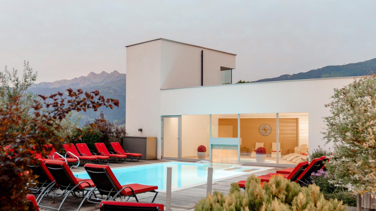 Ein stilvoll gestalteter Außenpool mit eleganten roten Liegestühlen lädt zum Entspannen ein. Im Hintergrund beeindruckt die alpine Berglandschaft, während das moderne Gebäude mit großen Glasfronten für eine helle und einladende Atmosphäre sorgt. Ideal für erholsame Stunden im Freien.