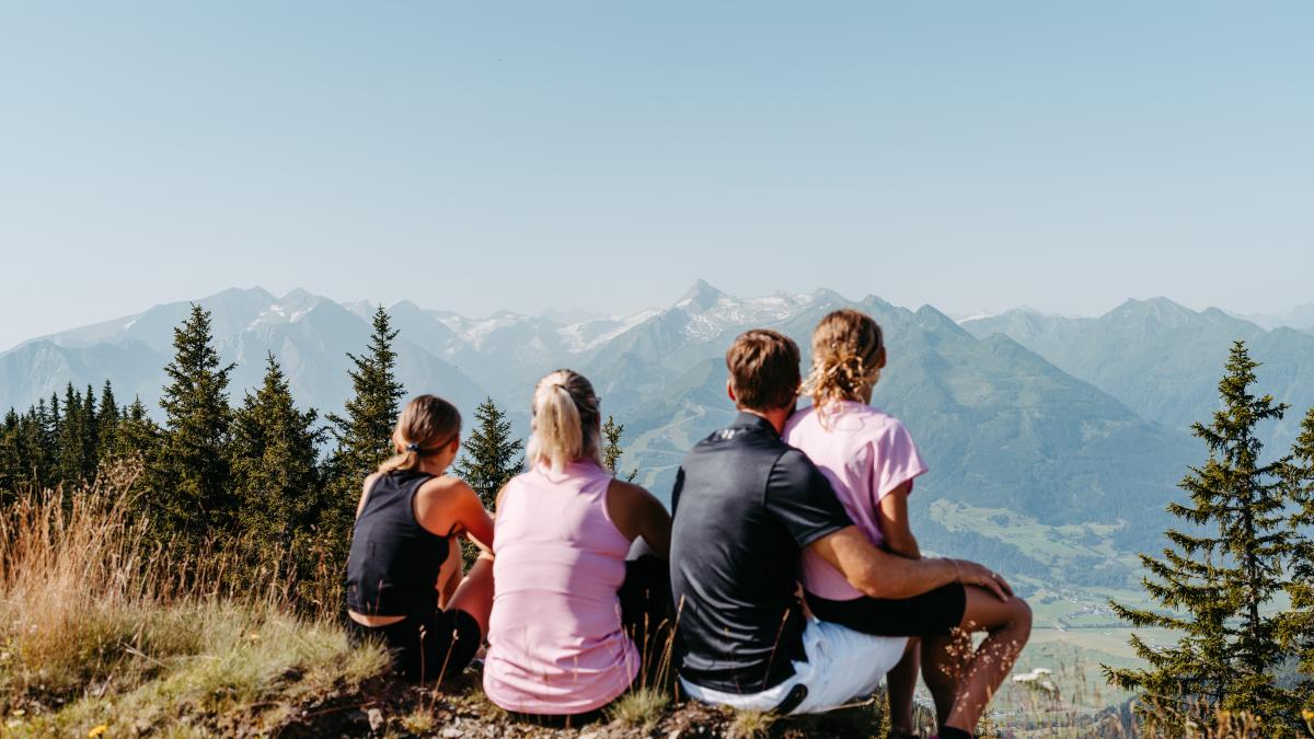 Vier Personen genie&szlig;en gemeinsam eine Pause auf einem Berggipfel und blicken auf die beeindruckenden Alpen. Umgeben von gr&uuml;nen B&auml;umen und sonniger Natur vermittelt das Bild Entspannung, Abenteuerlust und famili&auml;ren Zusammenhalt in malerischer Berglandschaft.