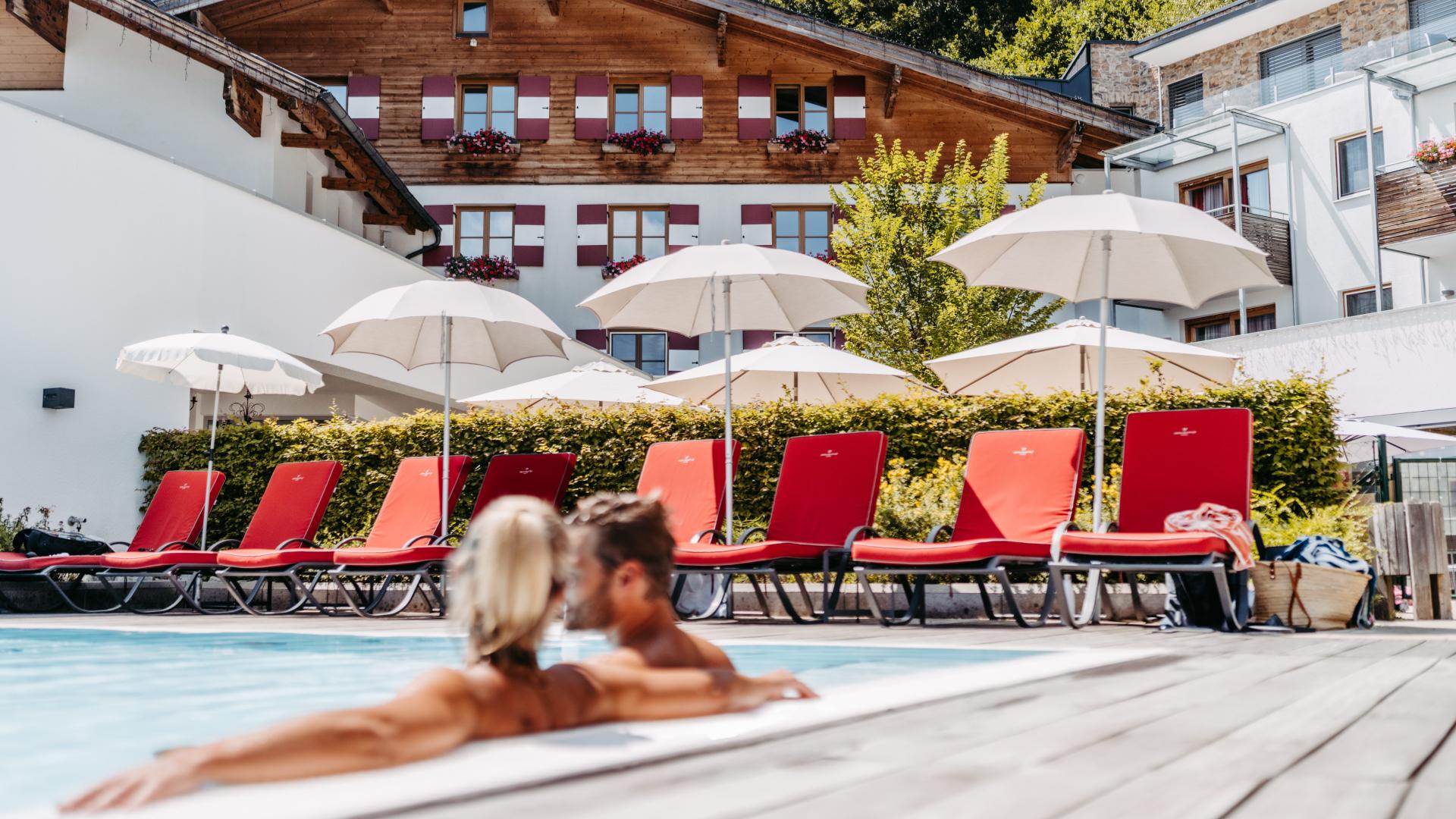 Ein Paar genießt einen sonnigen Tag im beheizten Außenpool eines gehobenen Resorts. Im Hintergrund sorgen rote Liegestühle und weiße Sonnenschirme vor einer charmanten Alpenkulisse für Urlaubsatmosphäre und ein Gefühl von Erholung und Wohlbefinden.
