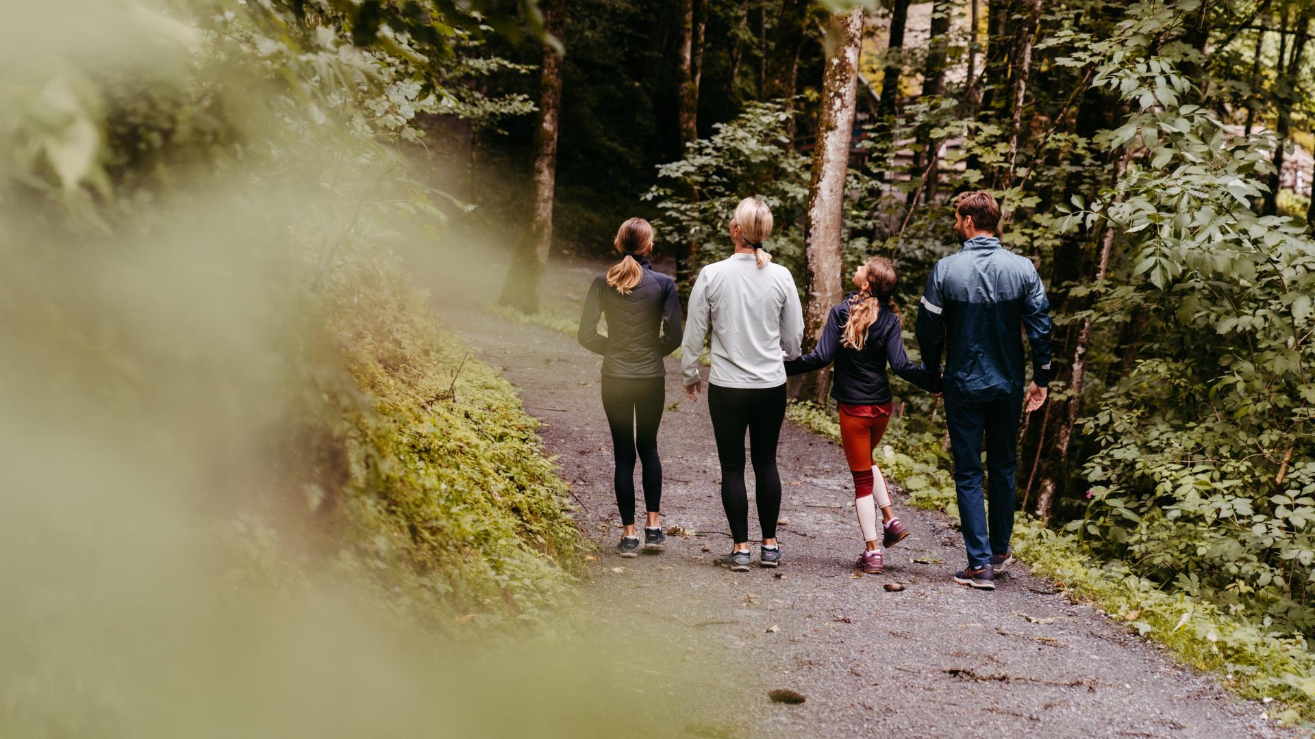 Eine Familie spaziert gemeinsam auf einem naturbelassenen Waldweg, umgeben von dichtem Gr&uuml;n und hohen B&auml;umen. Die entspannte Atmosph&auml;re l&auml;dt zu gemeinsamen Erlebnissen in der Natur ein. Sanftes Licht unterstreicht die friedliche Stimmung dieser Wanderung im Freien.