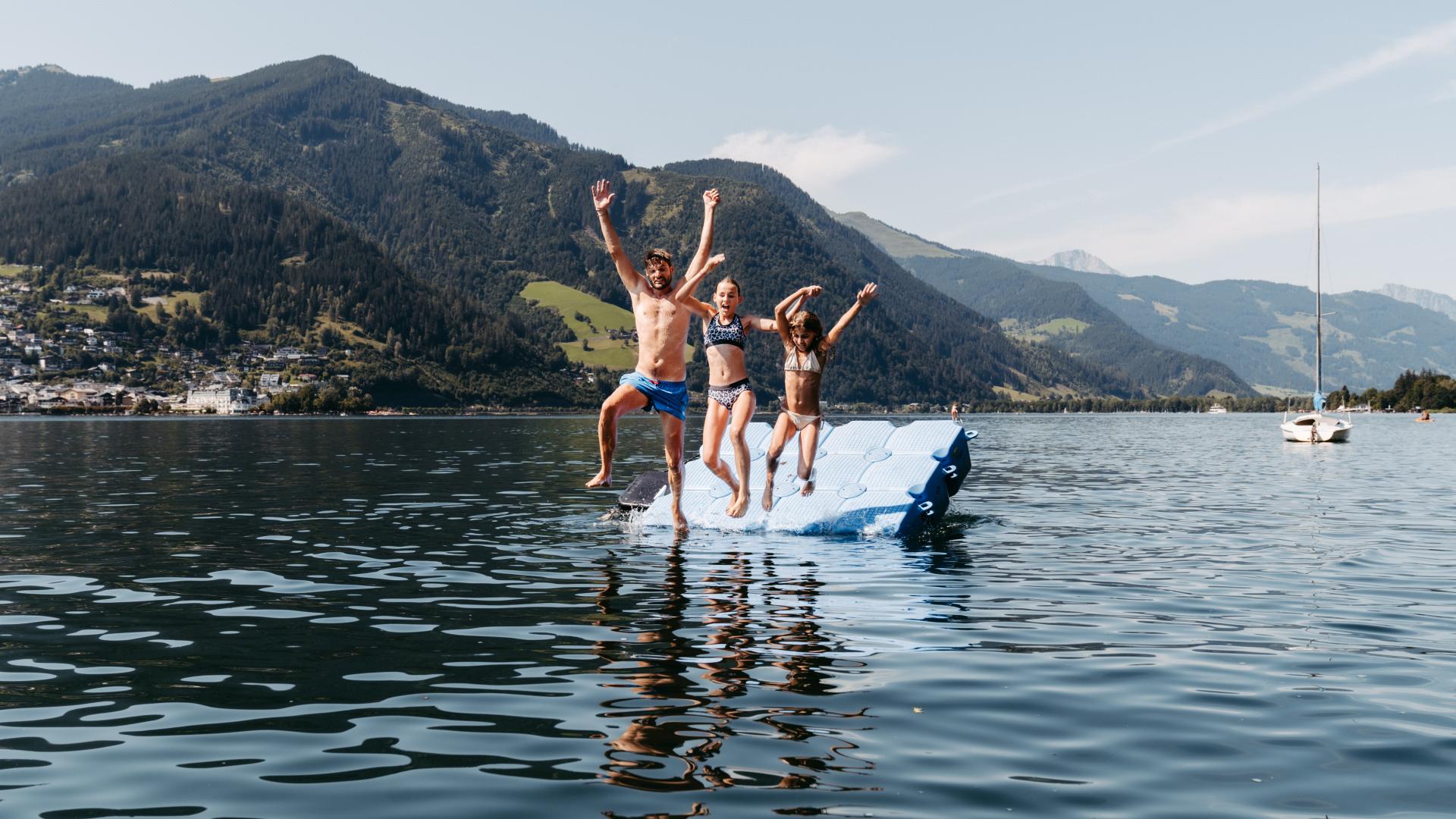 Eine fr&ouml;hliche Familie springt gemeinsam von einer Schwimminsel ins klare Wasser eines Alpensees. Im Hintergrund ragen bewaldete Berge auf und ein Segelboot liegt ruhig vor Anker. Die Szene strahlt Lebensfreude und Sommerurlaubsstimmung in idyllischer Naturkulisse aus.