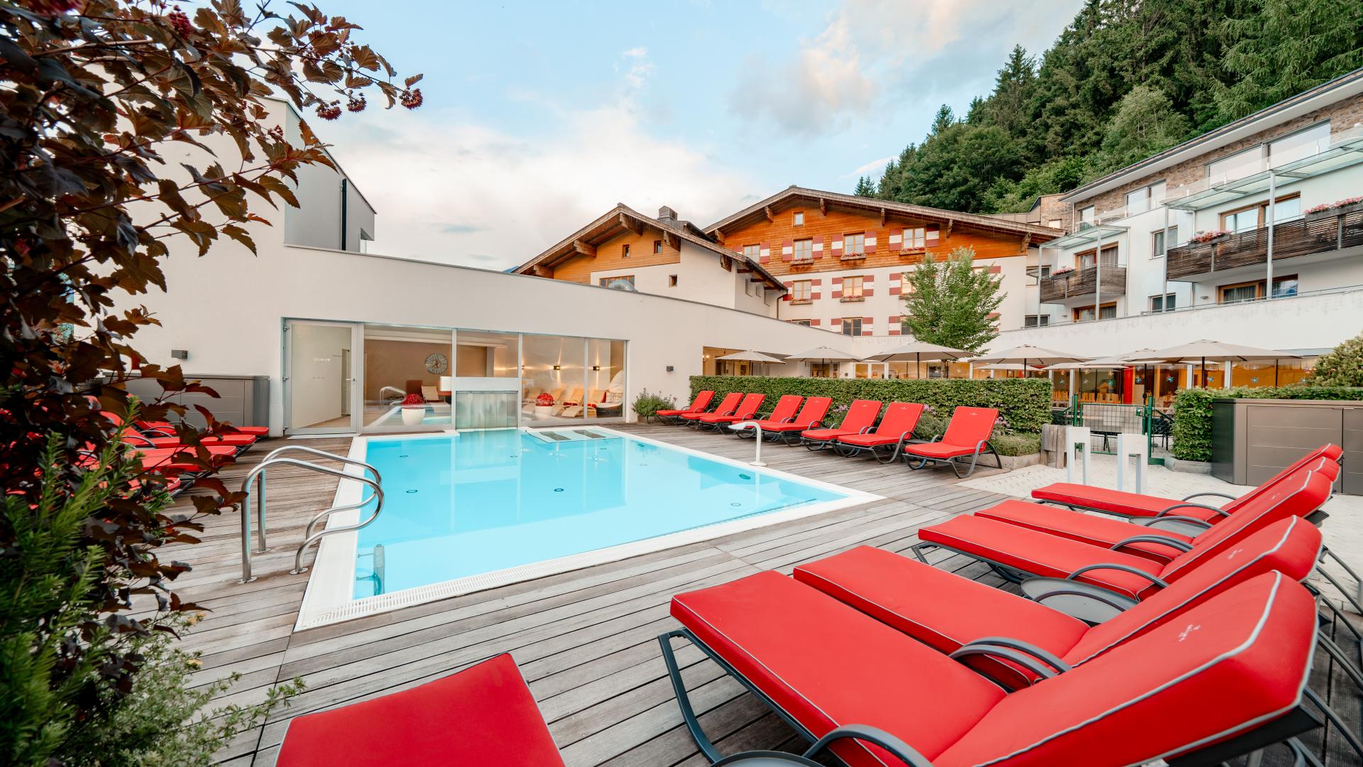 Moderner Außenpool inmitten einer stilvollen Holzterrasse mit bequemen roten Liegestühlen, eingerahmt von alpiner Natur. Im Hintergrund sind gemütliche Hotelgebäude und grüne Wälder zu sehen. Perfekt zum Entspannen und Genießen der ruhigen Atmosphäre im amiamo Familienhotel.