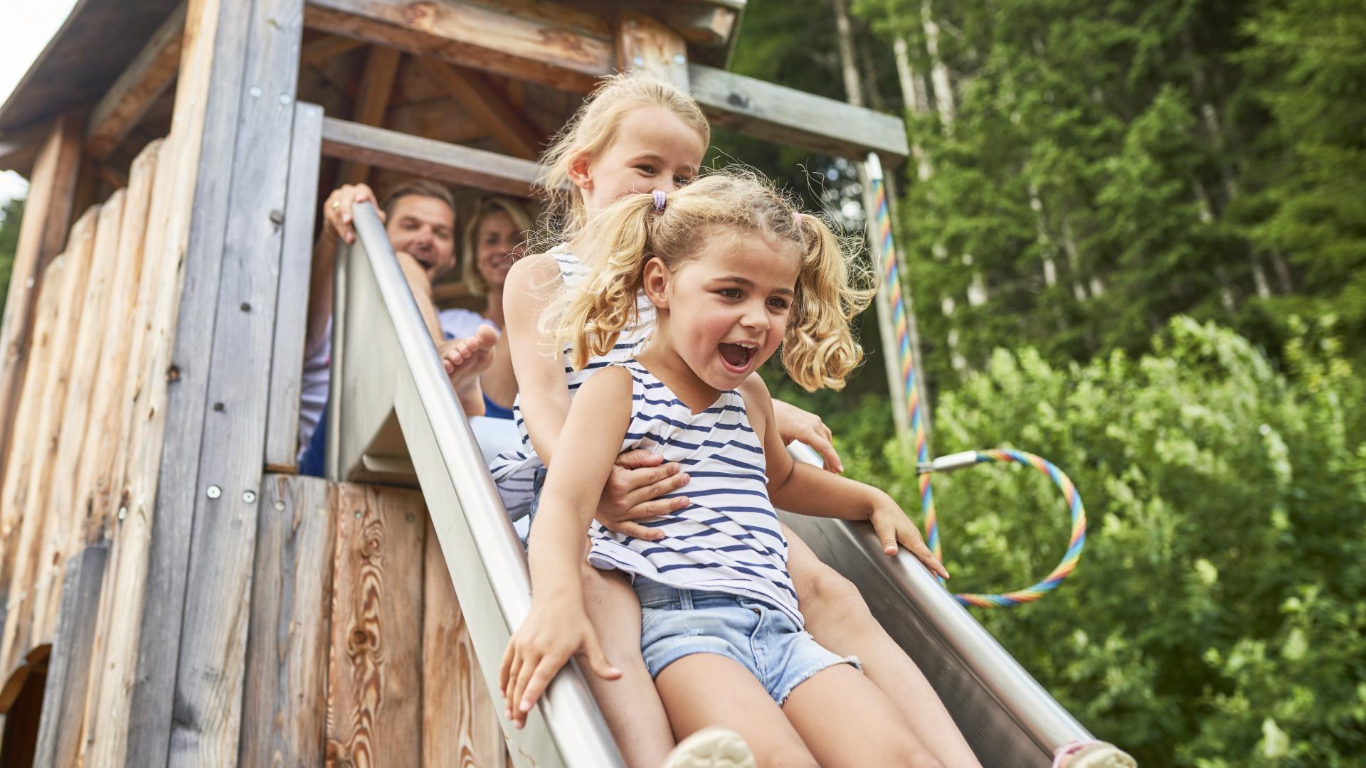 Mehrere fröhliche Kinder rutschen gemeinsam eine Holzrutsche im Grünen hinunter. Das Bild zeigt pure Lebensfreude und sommerlichen Spielspaß im Freien vor einem naturbelassenen Hintergrund mit Bäumen und Holzhaus. Ideal für Familienurlaube und Outdoor-Aktivitäten.