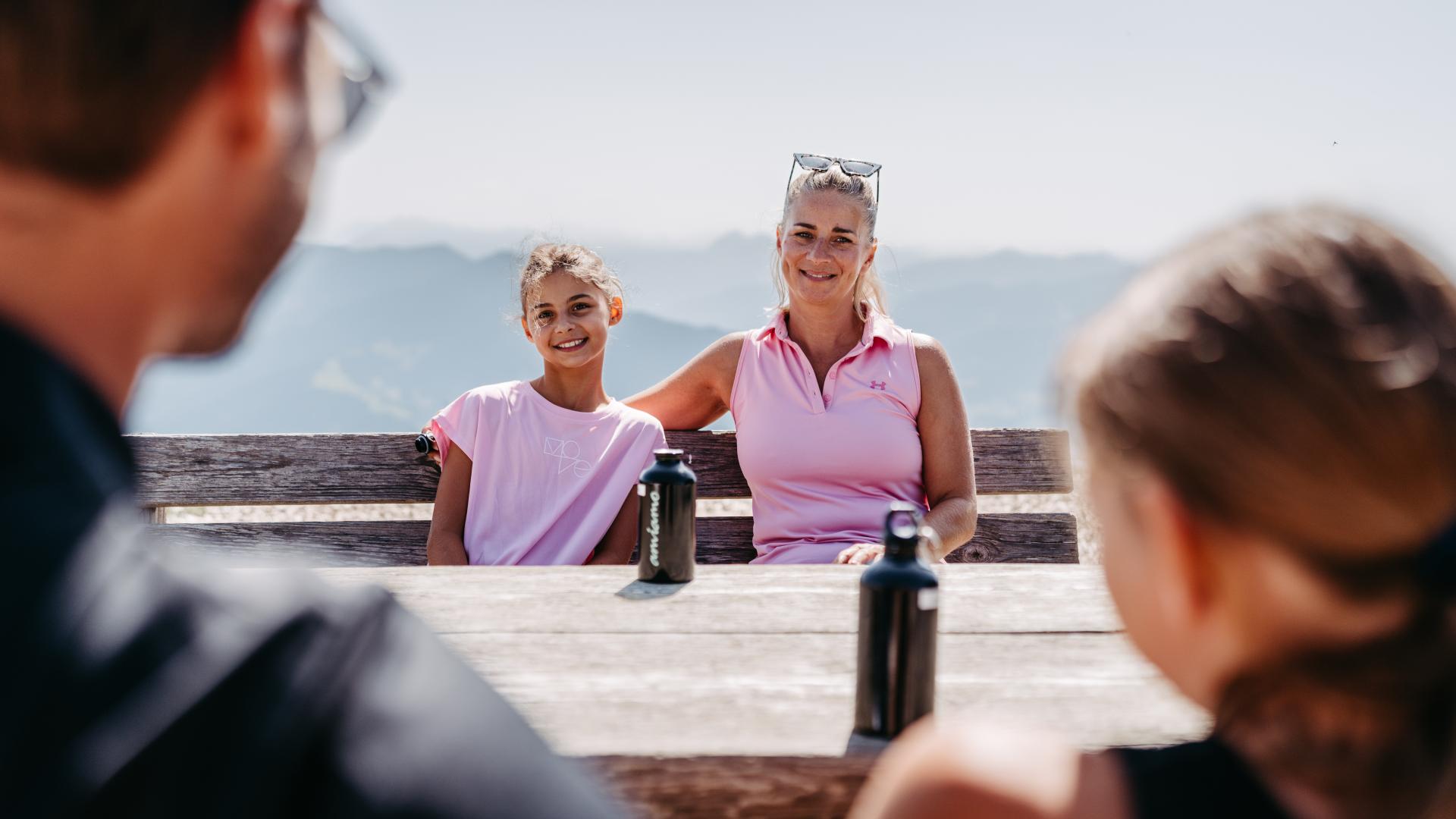 Eine lachende Mutter mit ihrem Kind genießt den Tag auf einer Berghütte, während Vater und Tochter ihnen gegenüber sitzen. Im Hintergrund erstreckt sich eine beeindruckende Alpenlandschaft. Wasserflaschen auf dem Holztisch runden die gemütliche Stimmung ab.