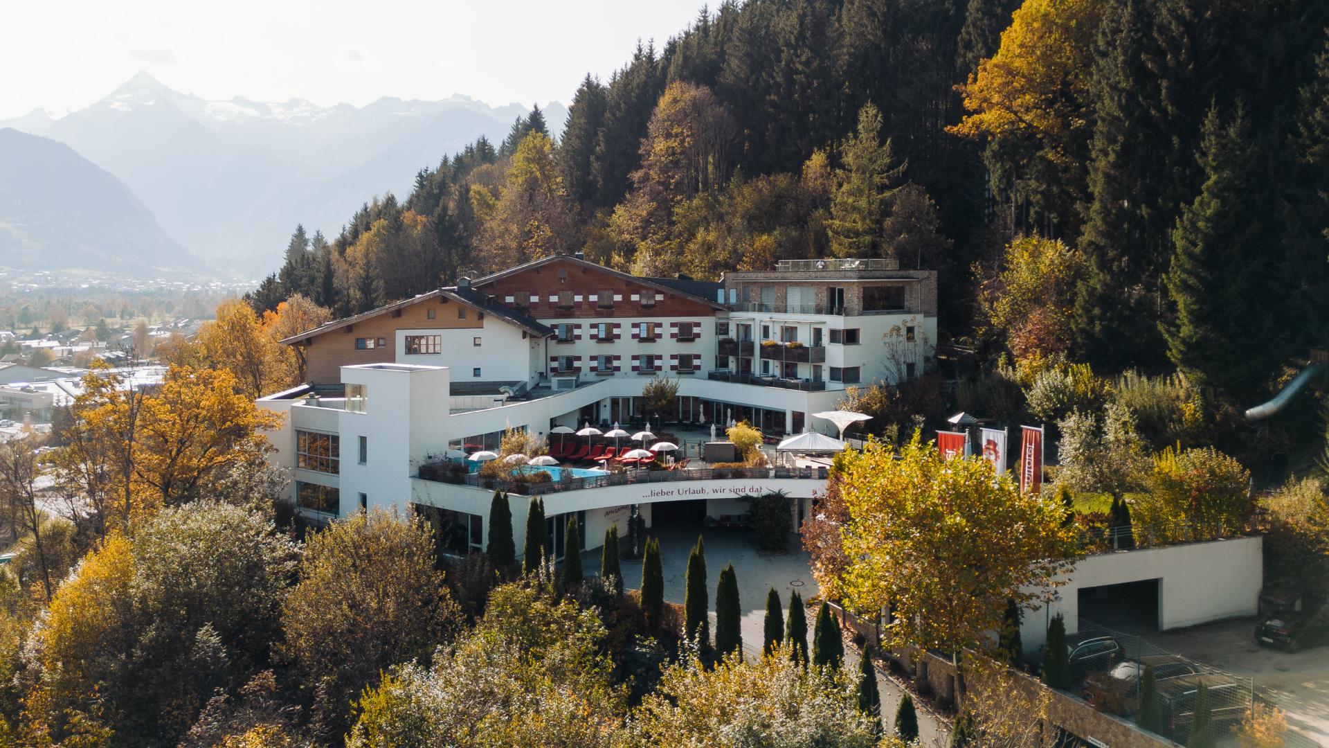 Das Bild zeigt das Hotel amiamo am Waldrand, eingebettet in eine herbstliche Alpenlandschaft mit goldenen und grünen Bäumen. Im Vordergrund ist eine sonnige Terrasse sichtbar, im Hintergrund ragen majestätische Berge auf. Die idyllische Lage bietet Ruhe und einen beeindruckenden Ausblick.