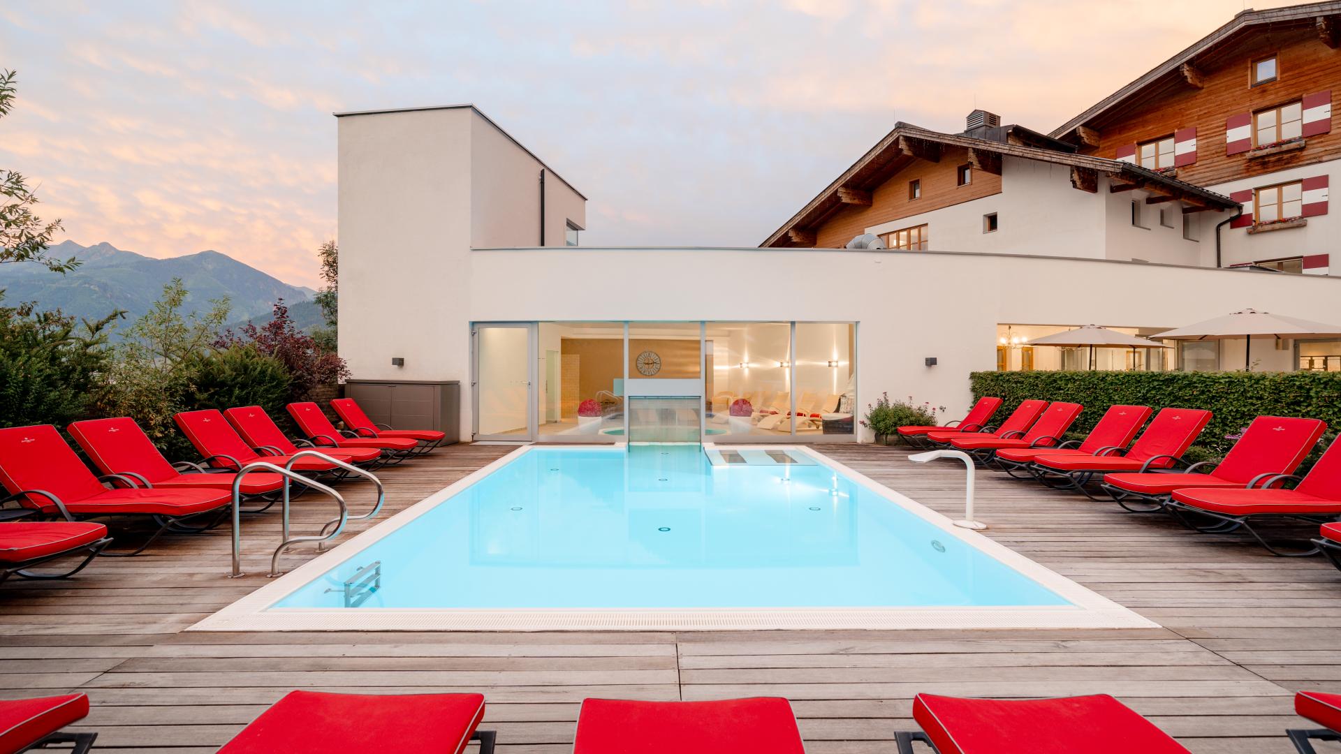 Modern outdoor pool with clean lines, surrounded by stylish red deckchairs on a wooden terrace. The pool area invites you to relax and offers an impressive view of the Alpine landscape. Ideal for individual time-outs and peaceful moments of well-being in a family-run hotel.