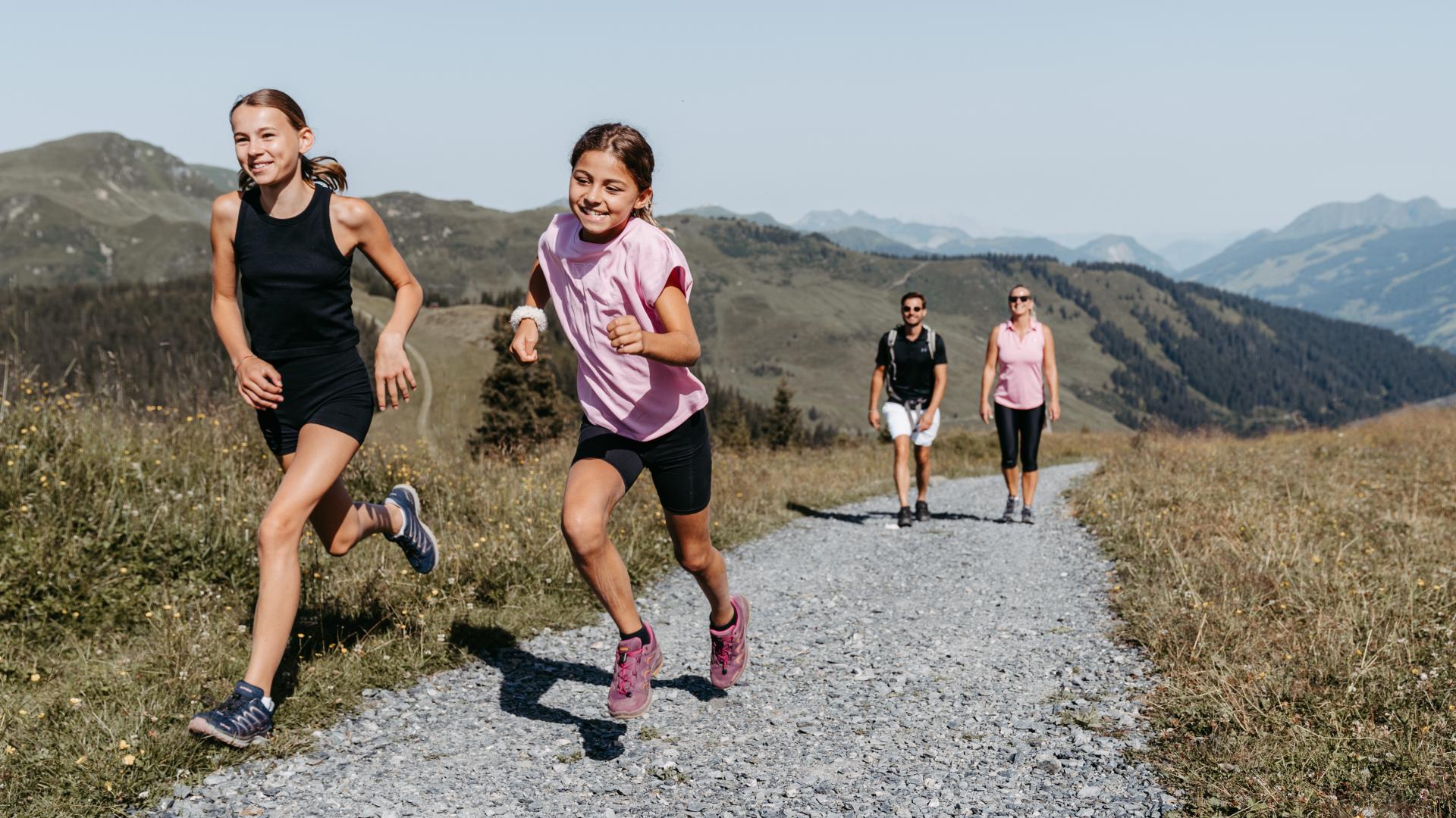 Zwei Kinder rennen fröhlich einen Schotterweg entlang, während im Hintergrund zwei Erwachsene gemütlich spazieren. Die Szene spielt inmitten einer idyllischen Alpenlandschaft, umgeben von grünen Hügeln und blauem Himmel – ideale Bedingungen für gemeinsame Familienaktivitäten in der Natur.