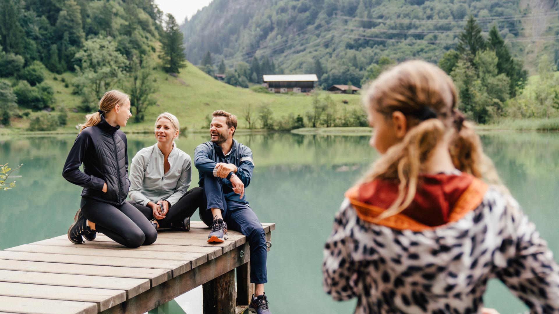 Eine fr&ouml;hliche Familie genie&szlig;t gemeinsame Zeit auf einem Steg an einem klaren Gebirgssee, umgeben von gr&uuml;ner Natur und Bergen. Im Vordergrund steht ein Kind, im Hintergrund entspannen die Eltern und ein weiteres Kind am Ufer. Perfekt f&uuml;r naturnahe Erholung und unvergessliche Momente.
