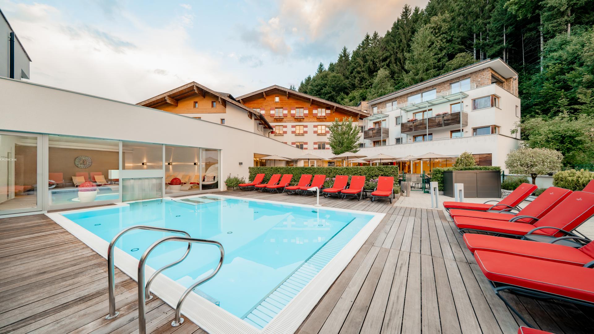 Moderner Außenpool mit breiter Holzterrasse und stilvollen roten Liegestühlen im Vordergrund. Im Hintergrund befindet sich ein elegantes Hotelgebäude, umgeben von grünen Bäumen am Waldrand. Klare Linien und warme Farben schaffen eine entspannte Urlaubsatmosphäre.