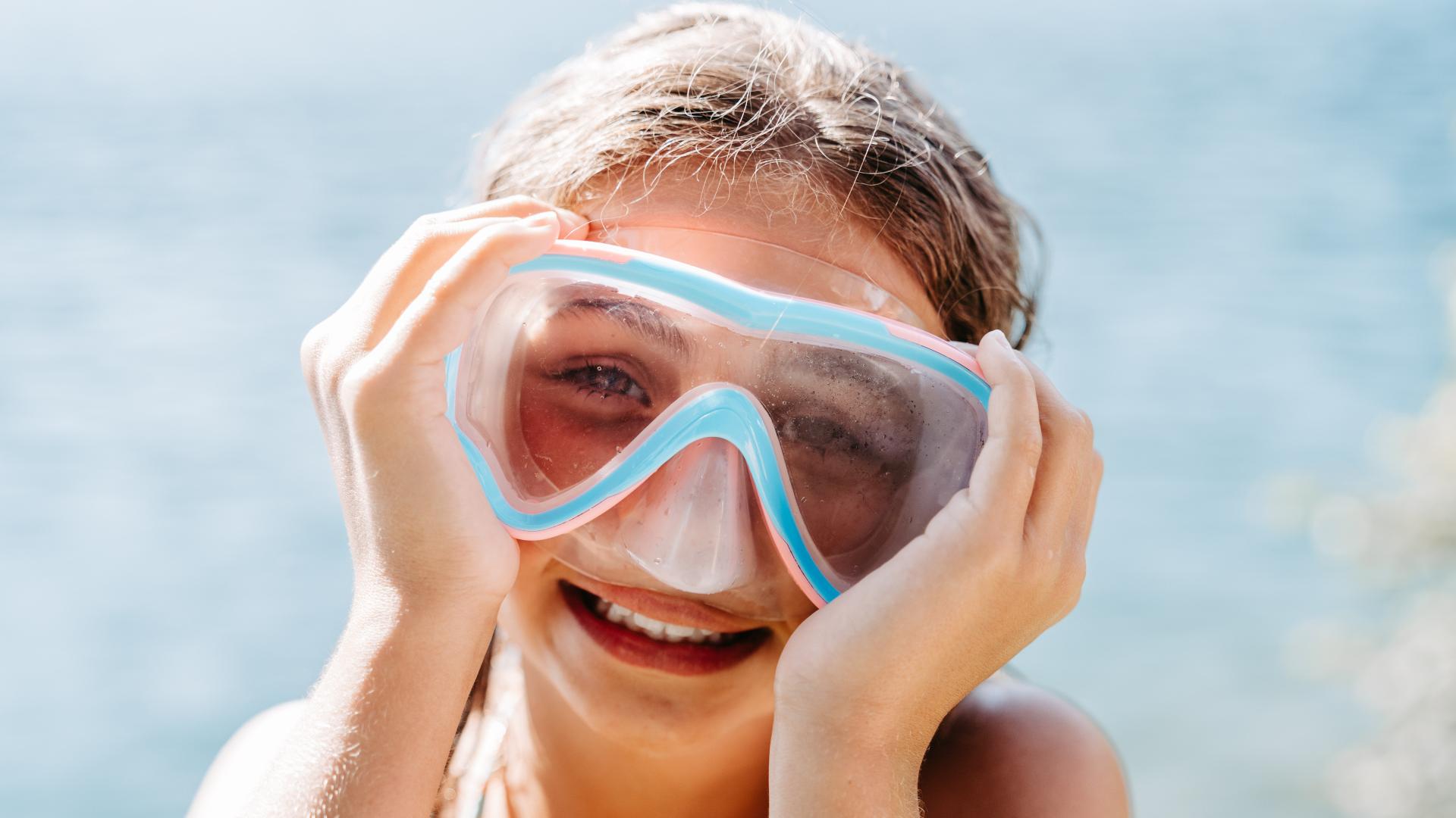 Ein lachendes Kind hält sich eine blaue Taucherbrille an das Gesicht und genießt den sonnigen Tag am Wasser. Im Hintergrund verschwimmt das ruhige Meer und vermittelt Urlaubsstimmung und Lebensfreude. Perfekt für sommerliche Familienmomente und unbeschwertes Badevergnügen.