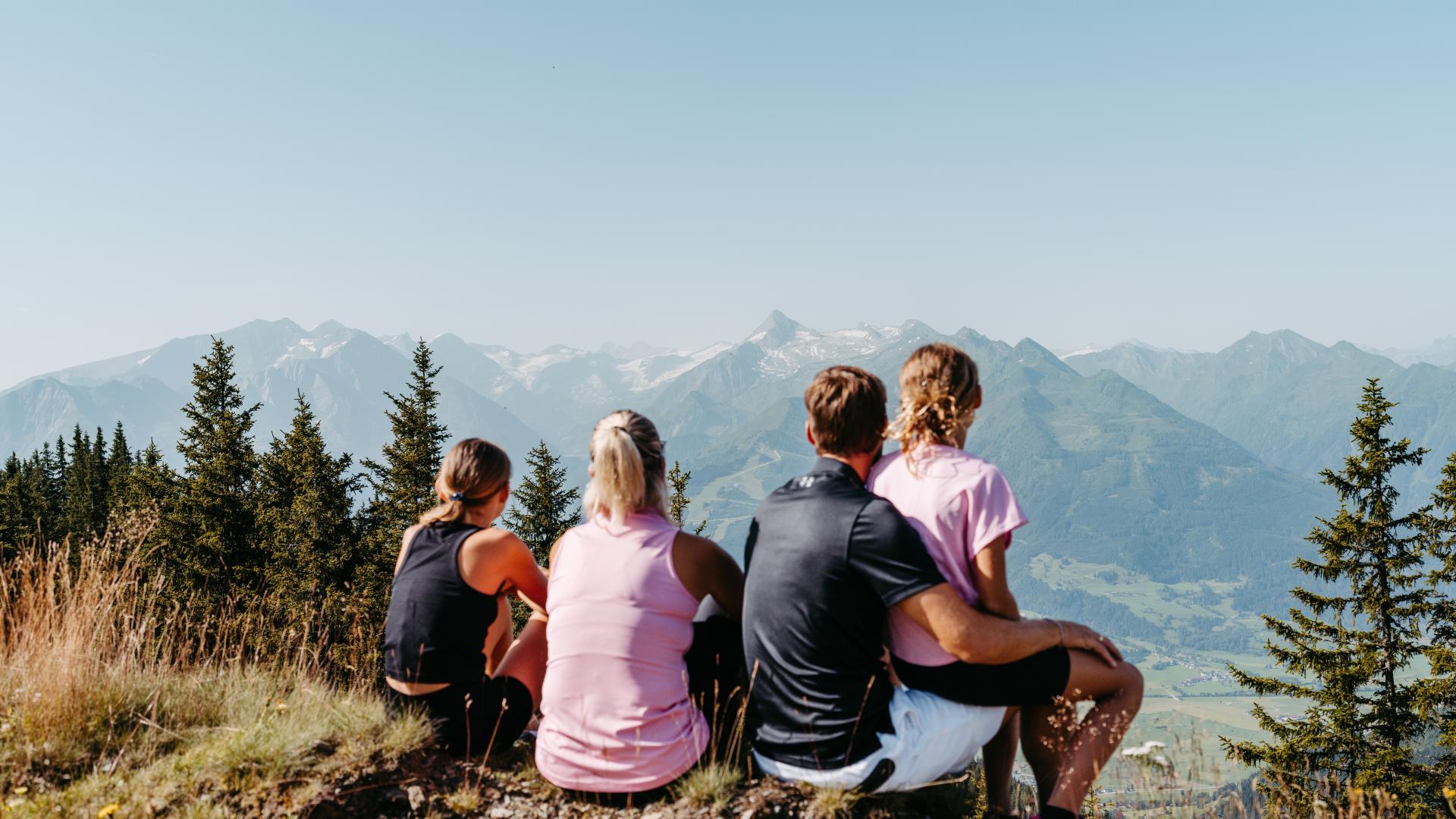 Vier Personen genießen gemeinsam eine Pause auf einem Berggipfel und blicken auf die beeindruckenden Alpen. Umgeben von grünen Bäumen und sonniger Natur vermittelt das Bild Entspannung, Abenteuerlust und familiären Zusammenhalt in malerischer Berglandschaft.
