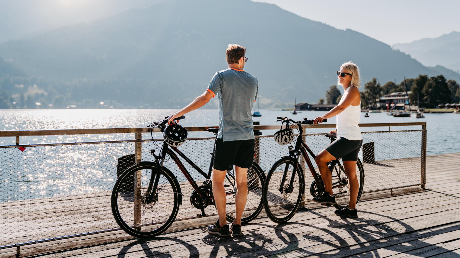 Zwei Menschen genießen mit ihren Fahrrädern eine entspannte Pause auf einem Holzsteg am sonnigen See. Im Hintergrund erheben sich sanfte Berge, das Wasser glitzert im Licht. Die Szene vermittelt pure Urlaubsfreude, Naturgenuss und aktive Erholung am Wasser.