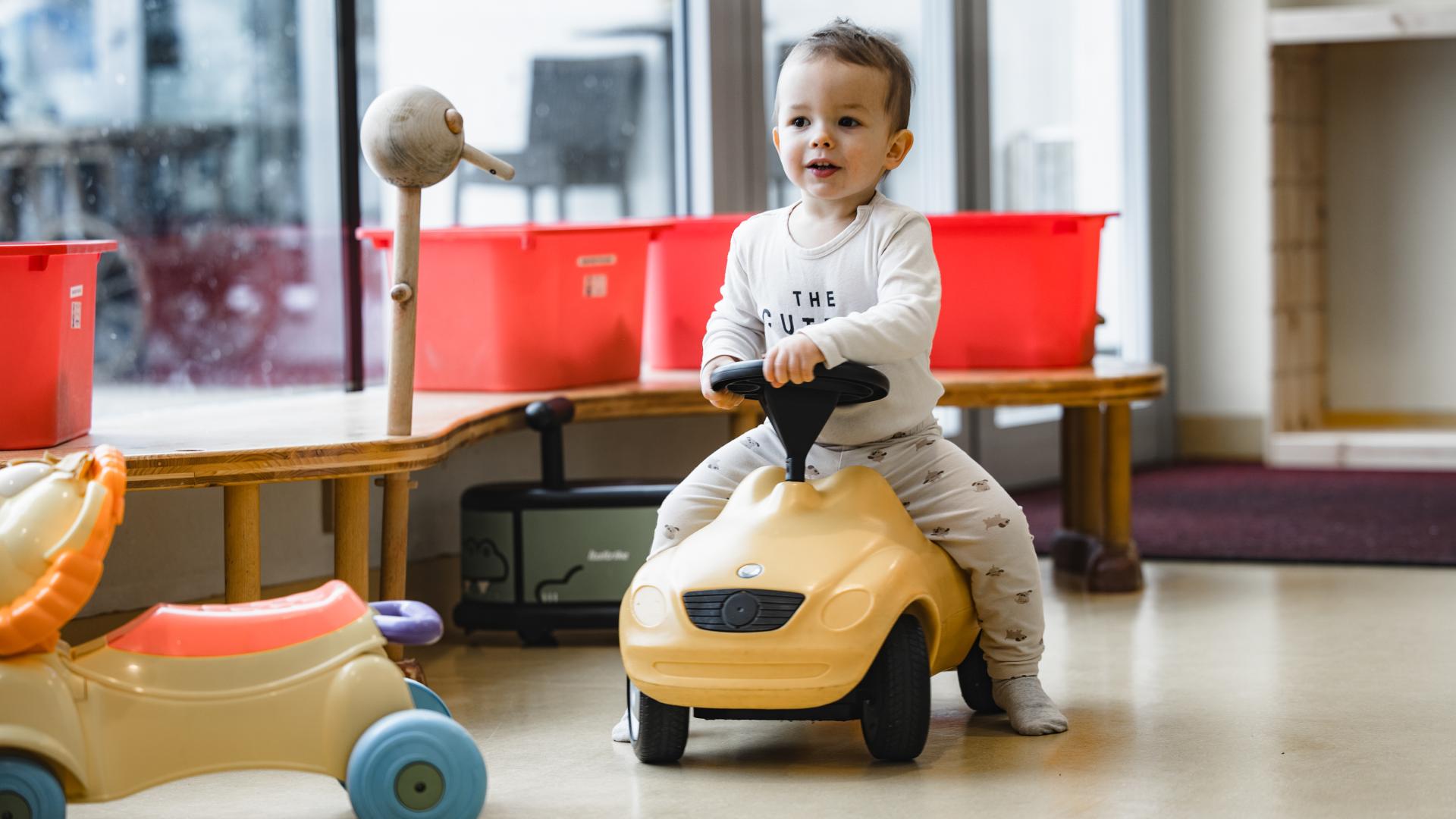 Ein fr&ouml;hliches Kleinkind f&auml;hrt auf einem gelben Rutschauto durch einen hellen, kinderfreundlichen Raum. Im Hintergrund stehen bunte Kisten und weiteres Spielzeug, was f&uuml;r eine spielerische, sichere Umgebung sorgt. Die Szene vermittelt Freude und Bewegungsfreiheit.
