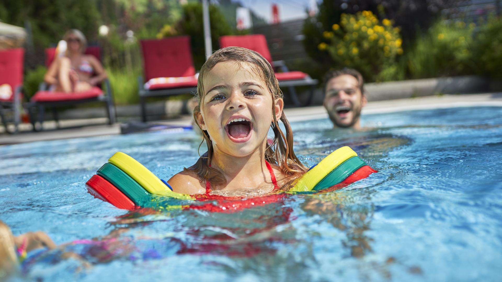 Ein lachendes Mädchen mit bunten Schwimmflügeln genießt das Baden im Außenpool, begleitet von einem Erwachsenen im Hintergrund. Sonnige Atmosphäre, rote Liegestühle und grüne Natur verstärken das sommerliche Urlaubsgefühl.