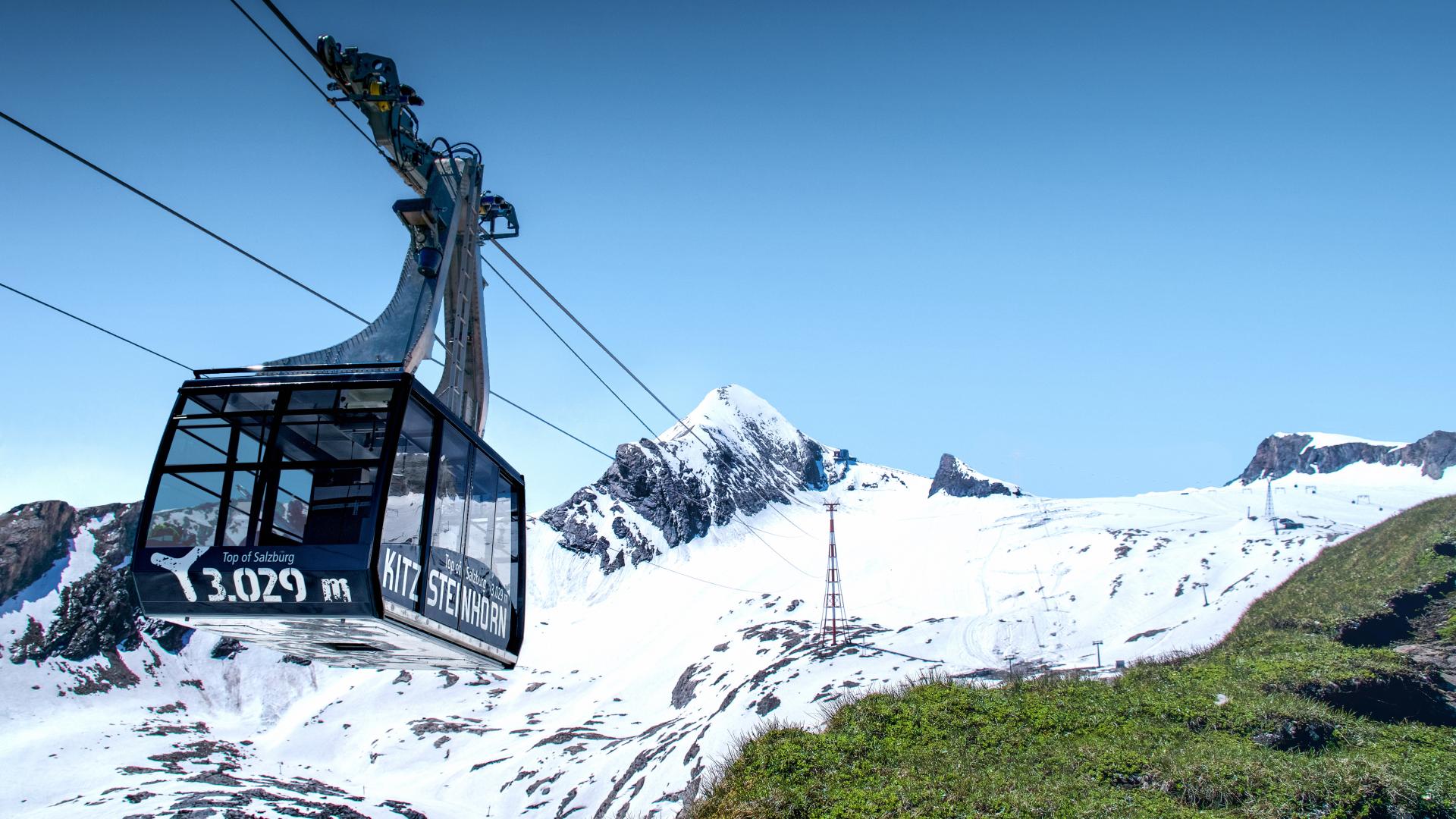 Eine moderne Seilbahngondel schwebt &uuml;ber die schneebedeckten H&auml;nge des Kitzsteinhorns in den Alpen. Der blaue Himmel und das markante Bergpanorama schaffen eine beeindruckende Kulisse. Die Gondel bietet einen Panoramablick auf das alpine Winterwunderland und gr&uuml;ne Almwiesen im Vordergrund.