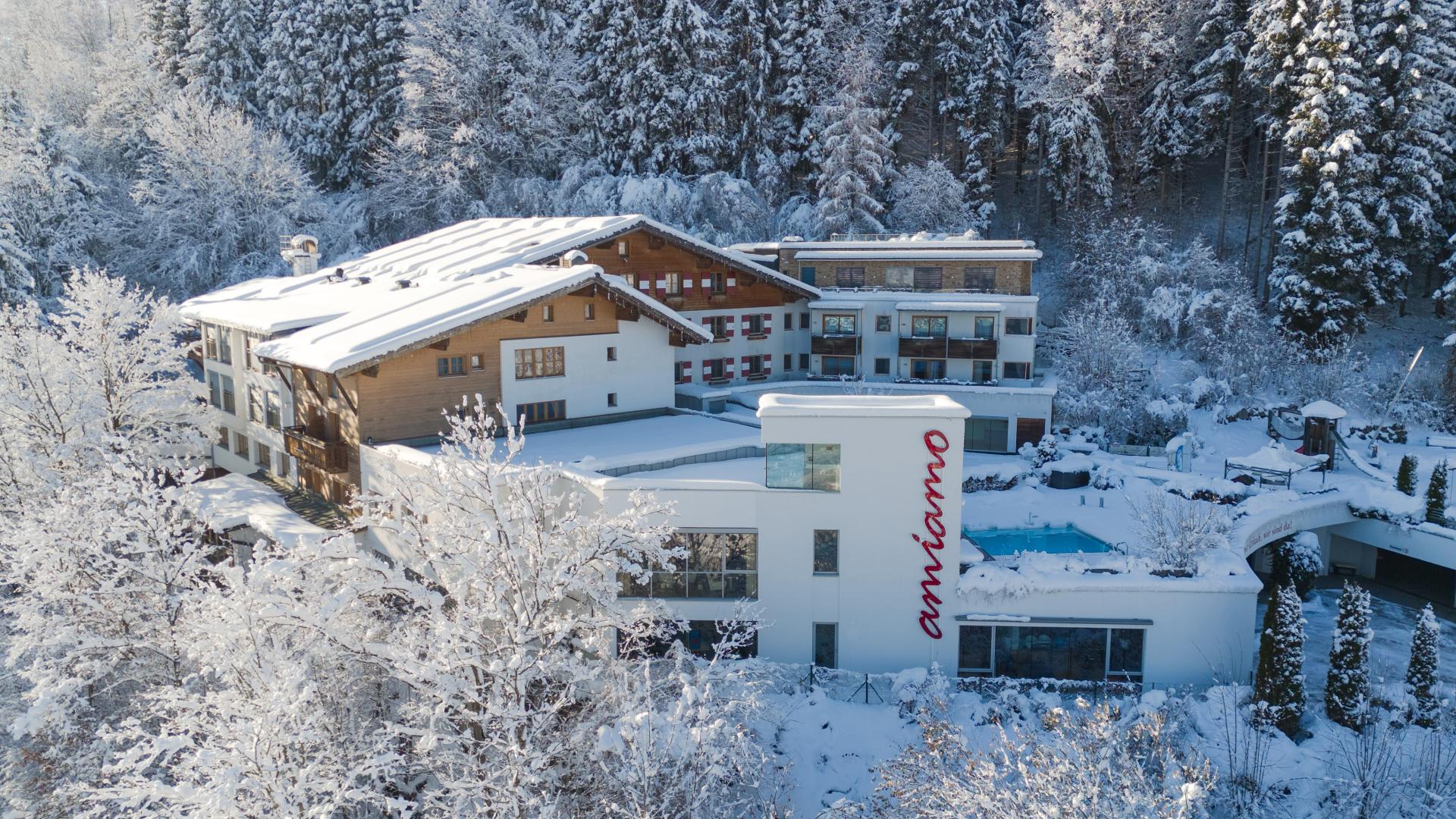 Familienhotel Amiamo im Salzburger Land im Winter &ndash; verschneite Hotelanlage mit traditionellem Holzchalet und modernem Wellnessbereich mit Au&szlig;enpool, eingebettet in eine malerische Winterlandschaft mit schneebedeckten Tannen