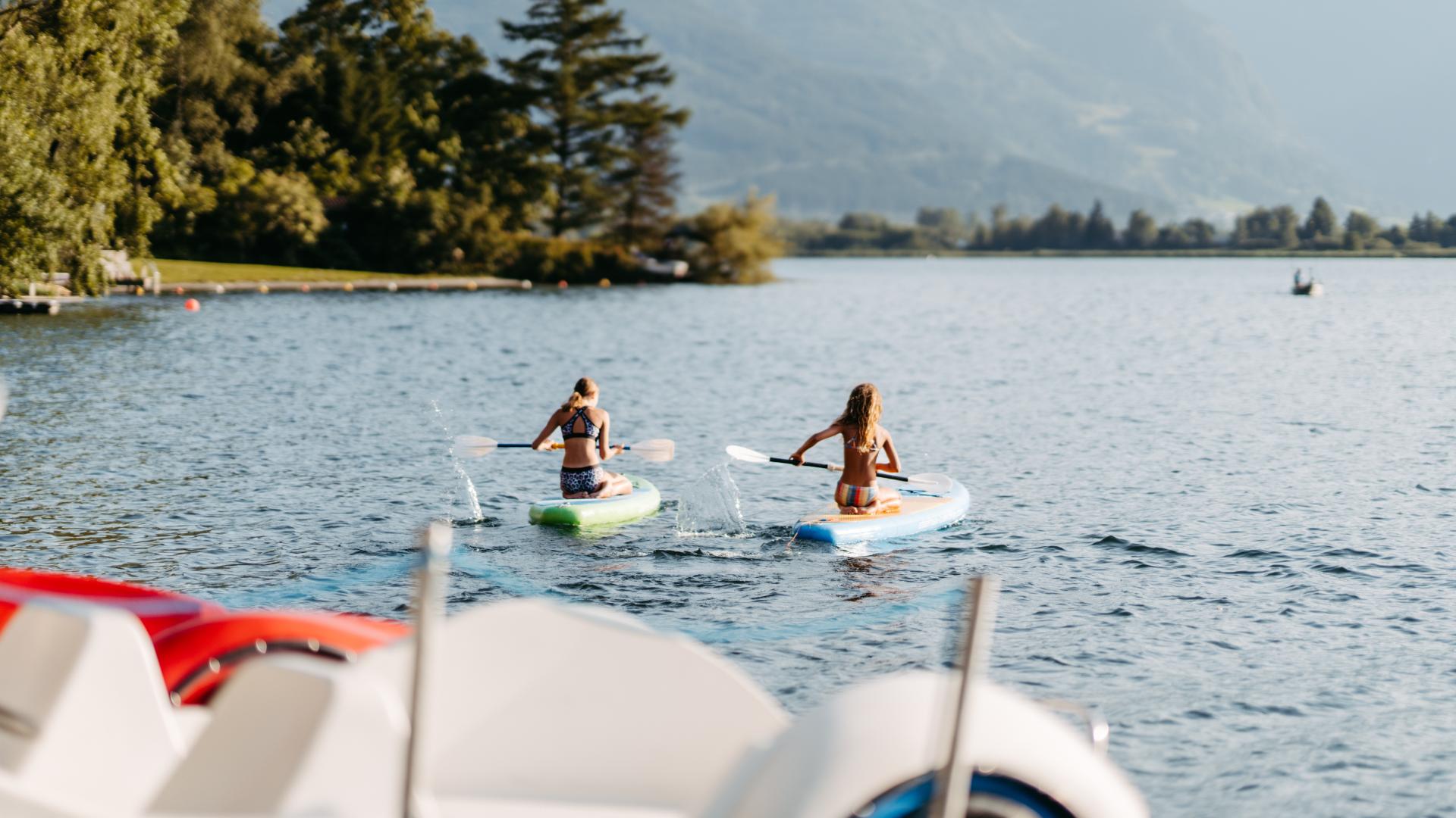 Zwei Personen genie&szlig;en eine entspannte Stand-Up-Paddling-Tour auf einem klaren See, umgeben von einer malerischen Bergkulisse und gr&uuml;nen B&auml;umen. Im Vordergrund sind Tretboote zu sehen, w&auml;hrend das ruhige Wasser und die idyllische Natur eine friedliche Atmosph&auml;re schaffen.