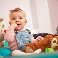 Ein lachendes Baby sitzt umgeben von bunten Kuscheltieren auf einer weichen Decke. Das Kind trägt einen rosa Body mit Jeans-Latzhose und ein Haarband, während es neugierig seine Hand im Mund hält. Die liebevolle Szene vermittelt Geborgenheit und kindliche Freude.