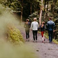 Eine Familie spaziert gemeinsam auf einem naturbelassenen Waldweg, umgeben von dichtem Gr&uuml;n und hohen B&auml;umen. Die entspannte Atmosph&auml;re l&auml;dt zu gemeinsamen Erlebnissen in der Natur ein. Sanftes Licht unterstreicht die friedliche Stimmung dieser Wanderung im Freien.