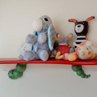 Verschiedene bunte Kuscheltiere, darunter ein grauer Elefant, ein Streifenhase und eine freundliche Raupe, sitzen ordentlich auf einem roten Wandregal. Die Szene wirkt einladend und kindgerecht und eignet sich perfekt für jedes Kinderzimmer.