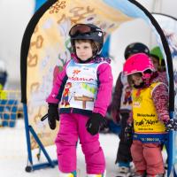 Mehrere Kinder nehmen an einem Skikurs teil und tragen bunte Skiausrüstung sowie Helme. Im Vordergrund steht ein Mädchen in pinker Skibekleidung, bereit zur Abfahrt. Im Hintergrund sind weitere Kinder unter einem farbenfrohen Torbogen zu sehen. Die winterliche Szene vermittelt Freude und Sicherheit beim Skifahren lernen.