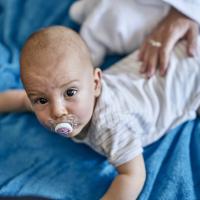 Ein Baby liegt auf dem Bauch auf einer weichen, blauen Decke und schaut neugierig nach oben. Das Kind tr&auml;gt einen Schnuller im Mund und helles, gestreiftes Body. Eine erwachsene Hand sorgt f&uuml;rs Wohlbefinden und vermittelt Geborgenheit. Die Szene wirkt liebevoll und geborgen.