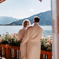 Ein Paar im Bademantel genie&szlig;t entspannt die Aussicht vom Balkon auf einen idyllischen See, umgeben von farbenfrohen Blumen. Im Hintergrund erheben sich majest&auml;tische Berge unter klarem Himmel. Die warme Atmosph&auml;re l&auml;dt zum Wohlf&uuml;hlen und Verweilen ein.