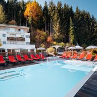 Moderner Au&szlig;enpool mit hellblauem Wasser, umgeben von roten Liegest&uuml;hlen und wei&szlig;en Sonnenschirmen vor einem stilvollen Alpenhotel. Die lebhaften Herbstfarben der umgebenden B&auml;ume sorgen f&uuml;r eine angenehme Wohlf&uuml;hl-Atmosph&auml;re und laden zum Entspannen ein. Ideal f&uuml;r erholsame Momente inmitten der Natur.