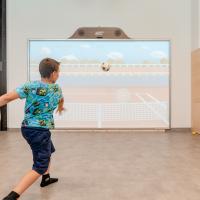 Ein Junge spielt Fu&szlig;ball an einer interaktiven Medienwand in einem Indoor-Spielbereich. Die Projektionsfl&auml;che zeigt ein virtuelles Spielfeld, das zum aktiven Mitmachen einl&auml;dt. Helle, freundliche Umgebung mit Kletterwand im Hintergrund und viel Bewegungsfreiheit f&uuml;r Kinder.