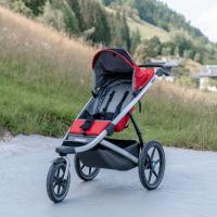 Ein moderner, rot-schwarzer Kinderwagen steht auf einem betonierten Weg, umgeben von grünen Wiesen und bewaldeten Hügeln. Die großen Räder und das stabile Gestell bieten idealen Komfort für Outdoor-Abenteuer. Im Hintergrund sind ländliche Gebäude und eine sanfte Berglandschaft zu sehen.