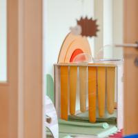 Ein freundlicher Kindergartenraum mit farbenfrohen Holzspielzeugen und weichen Matten. Durch eine offene T&uuml;r sieht man verspielte Dekorationen und einen Regenbogen aus Holz, die eine einladende und kreative Atmosph&auml;re f&uuml;r Kinder schaffen.