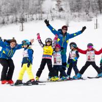 Fr&ouml;hliche Kindergruppe mit Skilehrern auf einer verschneiten Piste. Alle Kinder tragen bunte Skikleidung und Helme, w&auml;hrend die motivierten Skilehrer sie anleiten. Die winterliche Landschaft schafft eine freundliche und einladende Atmosph&auml;re f&uuml;r den gemeinsamen Lernspa&szlig; im Schnee.