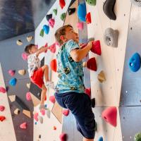 Zwei Kinder erklimmen eine farbenfrohe Boulderwand in einem hellen Indoor-Bereich. Die Wand ist mit bunten Griffen ausgestattet und f&ouml;rdert Bewegung, Geschicklichkeit und Teamgeist. Das Bild vermittelt Spa&szlig;, Konzentration und sportliche Aktivit&auml;t in sicherer Umgebung.