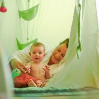 Ein Baby spielt fröhlich mit seiner Mutter in einem lichtdurchfluteten, grünen Spielzelt. Die Szene strahlt Geborgenheit und Freude aus, mit weichen Stoffelementen und liebevoller Atmosphäre – ideal für gemeinsame Entdeckungen und entspannte Momente.