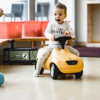 Ein fr&ouml;hliches Kleinkind sitzt auf einem gelben Rutschauto in einer hellen, freundlichen Spielumgebung. Im Hintergrund sind bunte Aufbewahrungsboxen und Fensterausblick zu sehen. Das Kind lacht begeistert und genie&szlig;t spielerisch seine Fahrt durch den Raum.