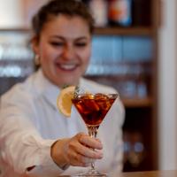 Eine l&auml;chelnde Barkeeperin reicht einen eleganten Cocktail im Martini-Glas &uuml;ber die Theke. Das Getr&auml;nk ist mit einer frischen Zitronenscheibe garniert und mit Eisw&uuml;rfeln gek&uuml;hlt. Im Hintergrund sind Flaschen und Gl&auml;ser in einer stilvollen Bar-Atmosph&auml;re zu sehen.