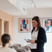 Eine Kellnerin lächelt herzlich und nimmt die Bestellung einer sitzenden Kundin in einem modernen Café entgegen. Im Hintergrund sorgen bunte Kunstwerke und gemütliche Sitzgelegenheiten für eine einladende Atmosphäre. Die freundliche Interaktion spiegelt Gastlichkeit und Wohlfühlambiente wider.