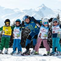 Eine fr&ouml;hliche Gruppe von Kindern posiert mit einem Skilehrer im Schnee, umgeben von verschneiten Bergen. Die Kinder tragen bunte Skiausr&uuml;stung und Helme, w&auml;hrend sie lachend miteinander stehen. Im Hintergrund erhebt sich ein beeindruckendes Alpenpanorama unter klarem Himmel.