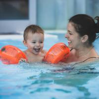 Eine Mutter genießt fröhlich das Schwimmen mit ihrem kleinen Kind, das orangefarbene Schwimmflügel trägt. Beide lächeln und verbringen gemeinsam unvergessliche Zeit im warmen Wasser eines Hallenbads. Die Szene vermittelt Freude, Sicherheit und Familienglück.