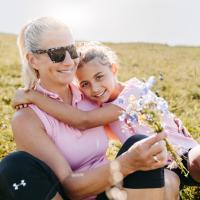 Eine Mutter und ihre Tochter genie&szlig;en gemeinsam einen sonnigen Tag auf einer Wiese. Die Mutter h&auml;lt einen bunten Wildblumenstrau&szlig; und das M&auml;dchen umarmt ihre Mutter liebevoll. Beide lachen gl&uuml;cklich, im Hintergrund erstreckt sich die gr&uuml;ne Landschaft unter klarem Himmel.