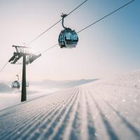 Eine moderne Seilbahn f&auml;hrt bei strahlendem Sonnenschein &uuml;ber eine frisch pr&auml;parierte Skipiste. Der glitzernde Schnee und die klaren Himmel vermitteln eine ruhige Winteratmosph&auml;re, ideal f&uuml;r Wintersportler in den Alpen. Die Bildkomposition betont Tiefe und Weite der verschneiten Landschaft.