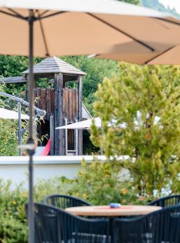 An inviting terrace with modern seating furniture and a large parasol in the foreground. In the background is a wooden playground with a slide, surrounded by green nature. Perfect for families who want to enjoy relaxing hours outdoors.