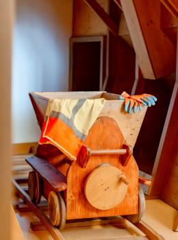 Ein liebevoll gefertigter Holz-Minenwagen steht auf Gleisen in einem kreativ gestalteten Kinderspielbereich. Ein farbenfroher Umhang und Handschuhe liegen &uuml;ber dem Wagen, was zum fantasievollen Spielen einl&auml;dt. Die warme Holzarchitektur sorgt f&uuml;r eine gem&uuml;tliche Atmosph&auml;re.