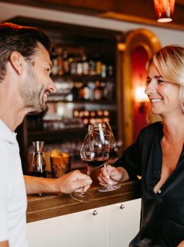 Ein entspanntes Paar genie&szlig;t ein Glas Rotwein an der stilvoll eingerichteten Bar. Im Hintergrund sind beleuchtete Flaschenregale sichtbar, die eine gem&uuml;tliche und einladende Atmosph&auml;re erzeugen. Die beiden Personen l&auml;cheln sich an und wirken gl&uuml;cklich, was dem Bild eine warme Stimmung verleiht.