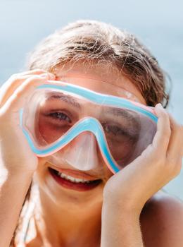 Ein lachendes Kind hält sich eine blaue Taucherbrille an das Gesicht und genießt den sonnigen Tag am Wasser. Im Hintergrund verschwimmt das ruhige Meer und vermittelt Urlaubsstimmung und Lebensfreude. Perfekt für sommerliche Familienmomente und unbeschwertes Badevergnügen.
