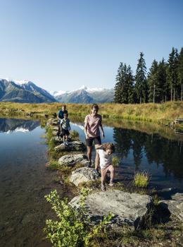Eine Familie &uuml;berquert gemeinsam einen idyllischen Bergsee auf Trittsteinen. Im Hintergrund erheben sich majest&auml;tische Alpen mit verschneiten Gipfeln und dichten W&auml;ldern. Das Sonnenlicht spiegelt sich im klaren Wasser, w&auml;hrend die Natur eine entspannte Urlaubsatmosph&auml;re bietet.