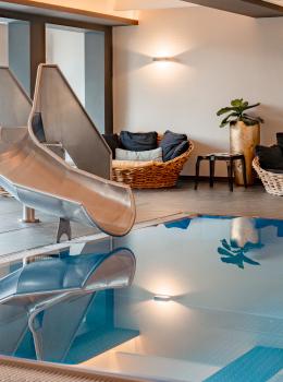 Ein stilvoller Innenpool mit einer gl&auml;nzenden Edelstahlrutsche sorgt f&uuml;r Spa&szlig; und Entspannung. Im Hintergrund laden gem&uuml;tliche Loungesessel und sanfte Beleuchtung zum Verweilen ein. Die klare Wasseroberfl&auml;che spiegelt die freundliche Atmosph&auml;re des Wellnessbereichs wider.