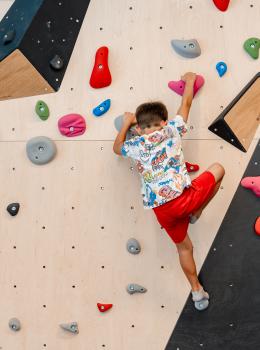 Ein Junge mit farbenfrohem T-Shirt und roten Shorts klettert an einer modernen Kletterwand mit bunten Griffen. Die Indoor-Kletterhalle bietet abwechslungsreiche Routen und sichere Bedingungen f&uuml;r junge Kletterbegeisterte. Die Szene vermittelt Bewegung, Konzentration und Freude am sportlichen Abenteuer.