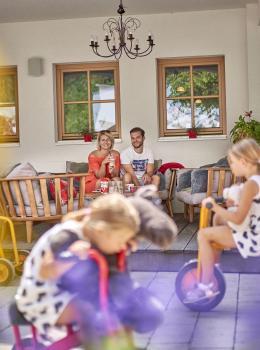 Auf einer sonnigen Terrasse entspannen zwei Erwachsene auf einer Holzbank, w&auml;hrend Kinder auf bunten Spielzeugen fr&ouml;hlich spielen. Die Szene vermittelt ein lebendiges, famili&auml;res Ambiente mit liebevollen Details und einer gem&uuml;tlichen, einladenden Atmosph&auml;re. Ideal f&uuml;r Familienurlaub und gemeinsame Momente.